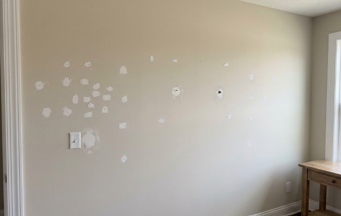Beige wall with dozens of white patched nail and anchor holes and a larger patch beside a light switch, before priming