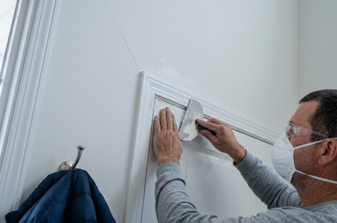 Painter in a mask and safety glasses working with a putty knife and mesh tape at the top corner of a door frame, with a diagonal crack running up the wall above