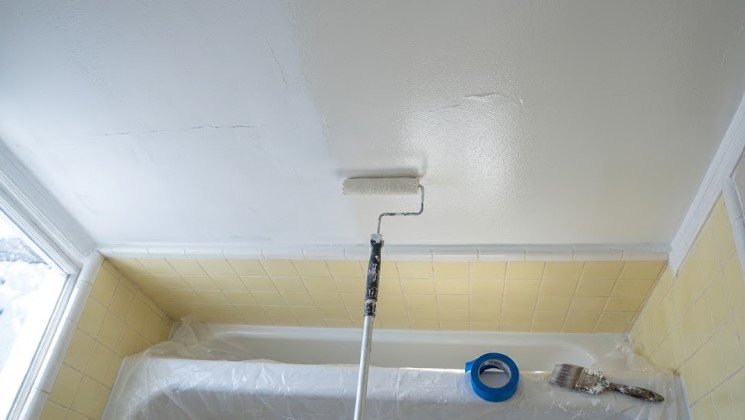 Paint roller applying white paint to a bathroom ceiling above a covered tub, with snow visible outside