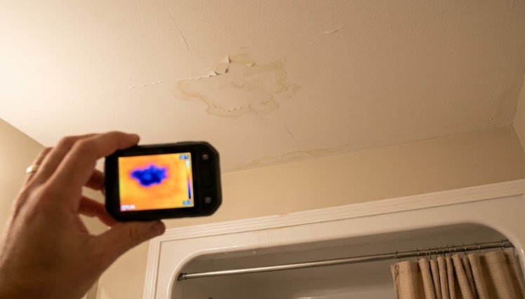 Hand holding a thermal camera showing a blue cold spot that matches peeling paint on a bathroom ceiling